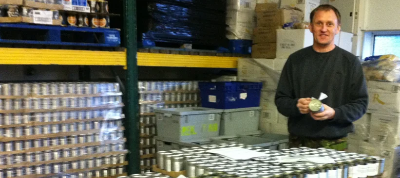 A Real Aid volunteer in the warehouse beside pallets stacked with tinned food